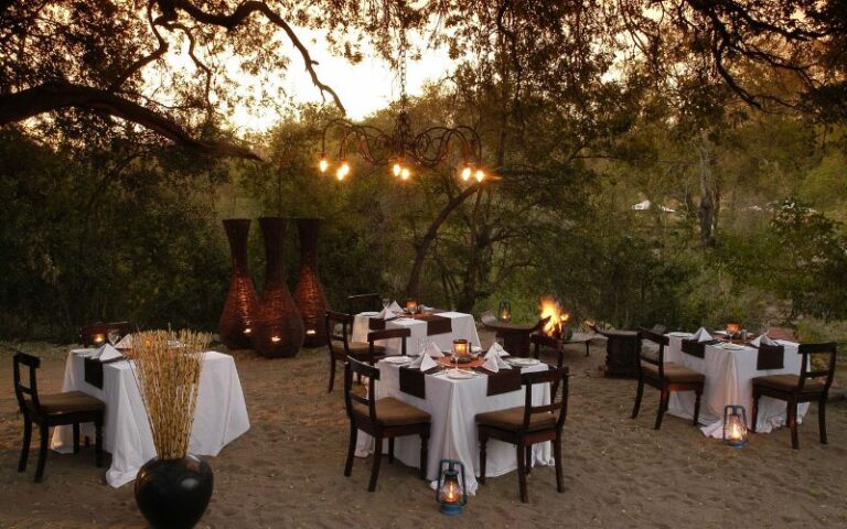 Elegant fireside safari dinner setup under the trees, with lantern lighting and tables arranged for outdoor dining in the African bush.