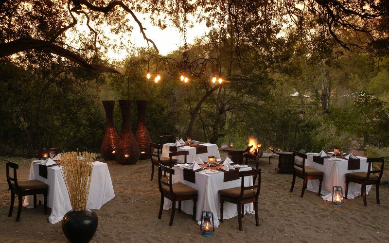 Elegant fireside safari dinner setup under the trees, with lantern lighting and tables arranged for outdoor dining in the African bush.