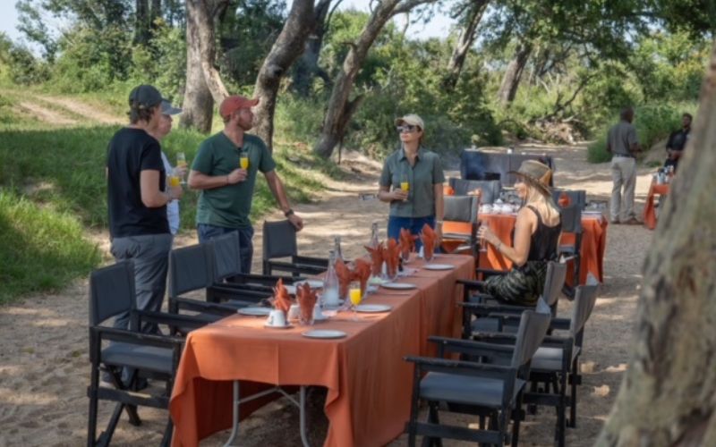 Guests enjoy a bush breakfast setup in the African wilderness with a beautifully laid table under shady trees.