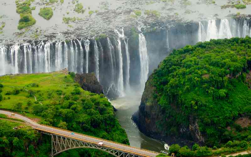 Victoria Falls bucket list experience is the breathtaking aerial view of Victoria Falls. This is where the mighty Zambezi River cascades over a dramatic gorge, creating mist and vibrant rainbows. A bridge stretches across the lush green landscape, connecting Zimbabwe and Zambia.