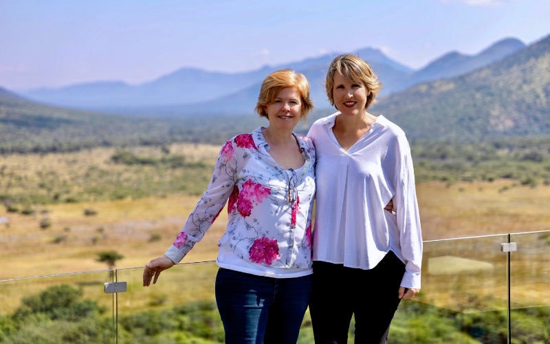 Two safari travel experts standing on a luxury South African lodge balcony overlooking the Big Five bushveld and mountain wilderness — symbolising women-led African safari experiences.